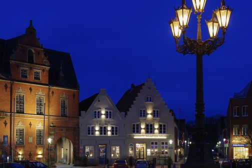 Historische Gebäude und eine verzierte Straßenlaterne erleuchten einen Platz bei Nacht. Die Fassade eines Hotels ist beleuchtet.