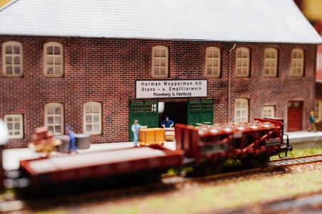Ein Miniaturmodell einer Fabrik mit der Aufschrift 'Herman Wupperman AG' und einer Modelleisenbahn im Vordergrund. Arbeiter sind beim Be- und Entladen zu sehen.