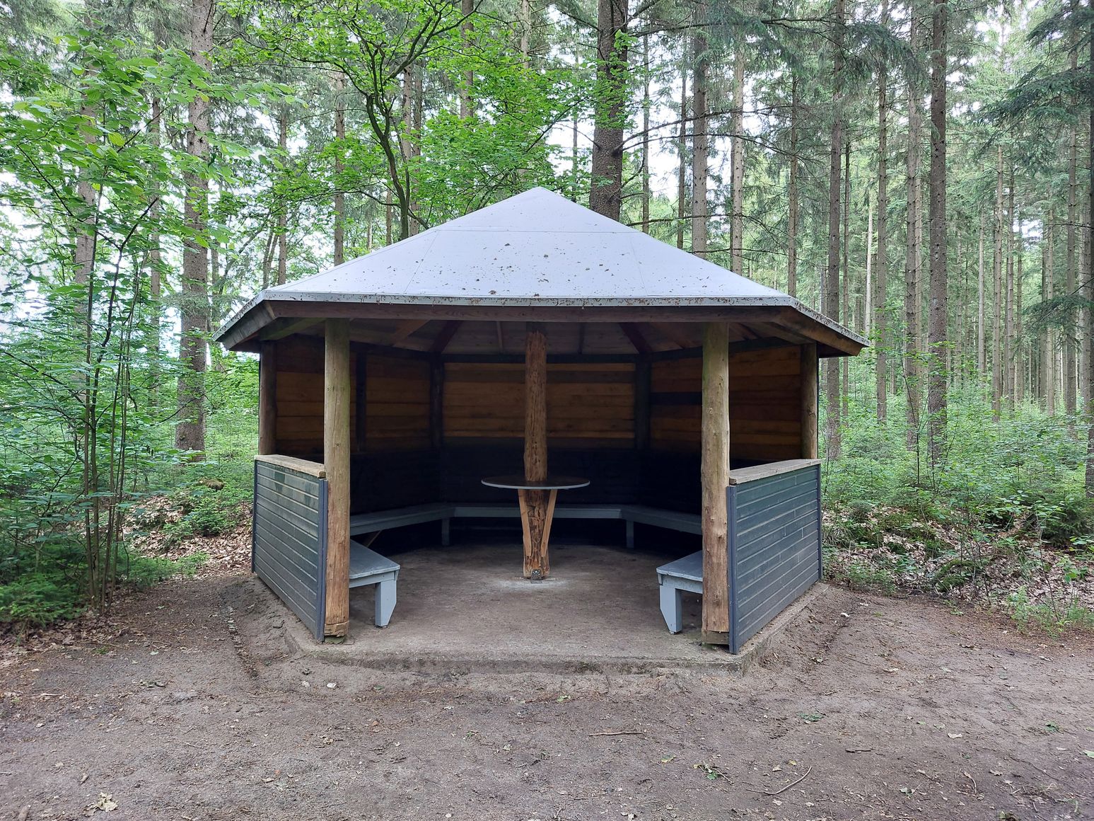 Offener Holzpavillon im Wald mit umlaufenden Bänken und Metall-Dach, umgeben von hohen Bäumen.