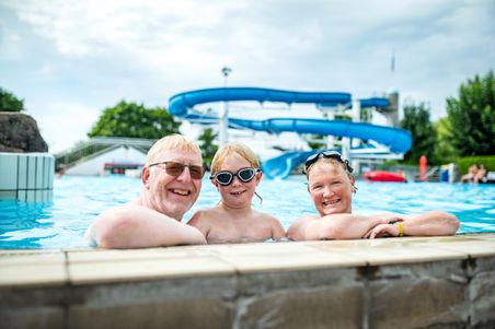 Eine Familie genießt einen Tag im Schwimmbad. Im Hintergrund ist eine große blaue Wasserrutsche zu sehen. Alle lächeln und tragen Schwimmbrillen.