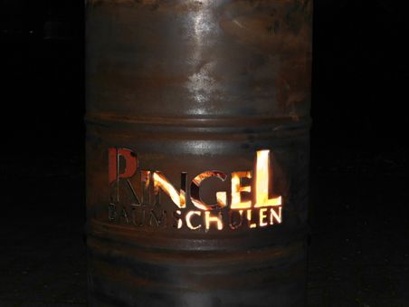 Metallfass mit beleuchtetem Schriftzug „Ringel Baumschulen“ vor schwarzem Hintergrund.