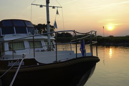 Ein Boot liegt bei Sonnenuntergang an einem ruhigen Flussufer vor Anker. Die Sonne spiegelt sich im Wasser und taucht die Szene in warmes Licht.