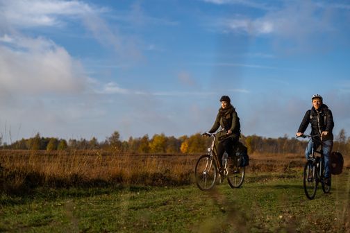 Zwei Radfahrer genießen eine Fahrt durch eine herbstliche Landschaft. Der Himmel ist blau mit einigen Wolken, und die Umgebung ist von braunen und grünen Tönen geprägt.