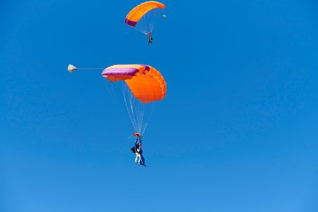 Zwei Fallschirmspringer mit orangefarbenen Fallschirmen schweben am klaren blauen Himmel. Einer der Springer ist im Tandemflug.