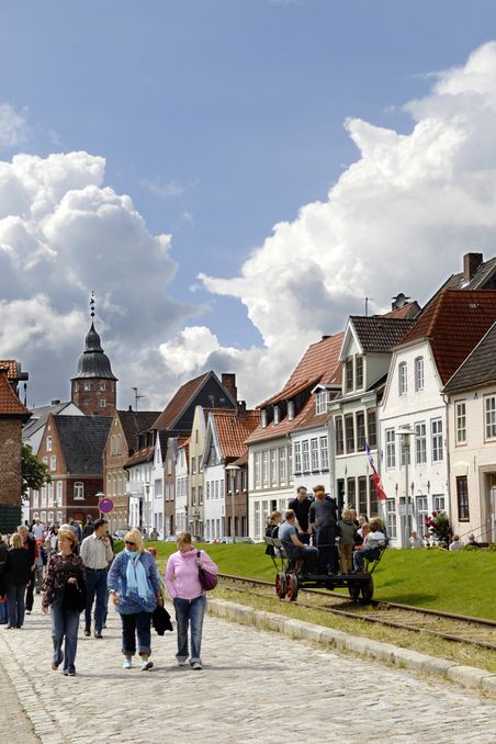 Menschen flanieren in Glückstadt entlang einer gepflasterten Straße, gesäumt von historischen Häusern. Ein Kirchturm erhebt sich im Hintergrund unter einem bewölkten Himmel.