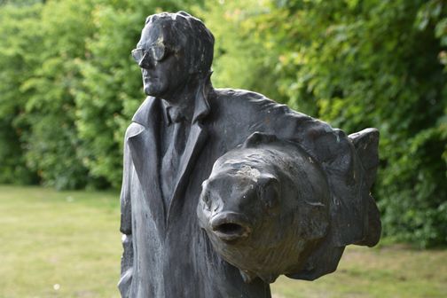 Bronzeskulptur im Skulpturengarten: ein Mann mit Brille neben einer abstrakten Kopf- oder Fischform vor grünem Grün.