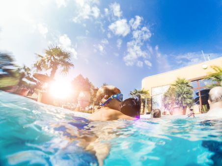 Menschen im Pool unter blauem Himmel, mit Palmen und starkem Sonnenlicht; ein Kind mit Schwimmbrille wird gehalten.