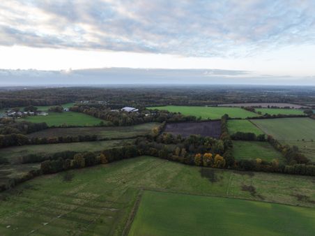 Luftaufnahme von einem Hof : grüne Felder, Wiesen und Waldlandschaft unter weitem, bewölktem Himmel.