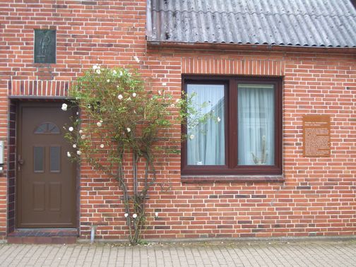 Backsteinfassade mit brauner Tür, Fenster und Kletterrose; rechts eine kleine Tafel an der Wand.