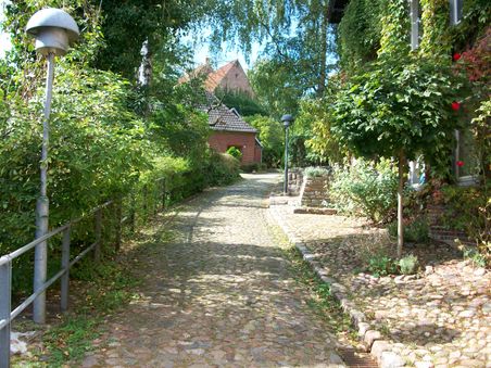 Gepflasterte Straße mit Bäumen und Sträuchern, die zu einem Backsteinhaus führt. Die Umgebung wirkt ruhig und grün.