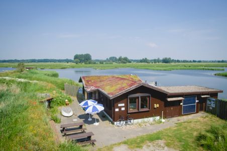 NABU Vogelstation Wedeler Marsch mit begrüntem Dach, Wasserflächen und weiter Marschlandschaft; davor NABU-Sonnenschirme, Tische und ein Fahrrad.