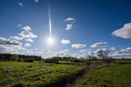 Grüne Felder unter einem strahlend blauen Himmel mit vereinzelten weißen Wolken und einer hellen Sonne.