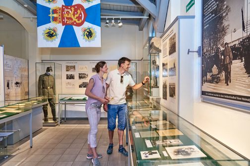 Ein Paar erkundet ein Museum mit historischen Exponaten, darunter Uniformen und Fotografien. Eine große Flagge hängt an der Decke.