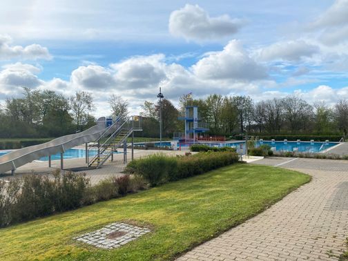 Freibad mit Rutsche, Sprungturm und Außenbecken unter wolkigem Himmel.