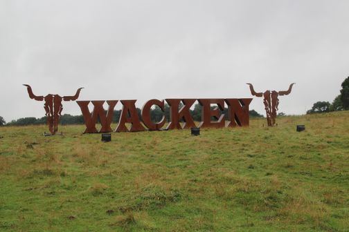 Der Schriftzug „Wacken“ steht auf einer grünen Wiese, links und rechts begleitet von zwei stilisierten Stierkopf-Skulpturen.