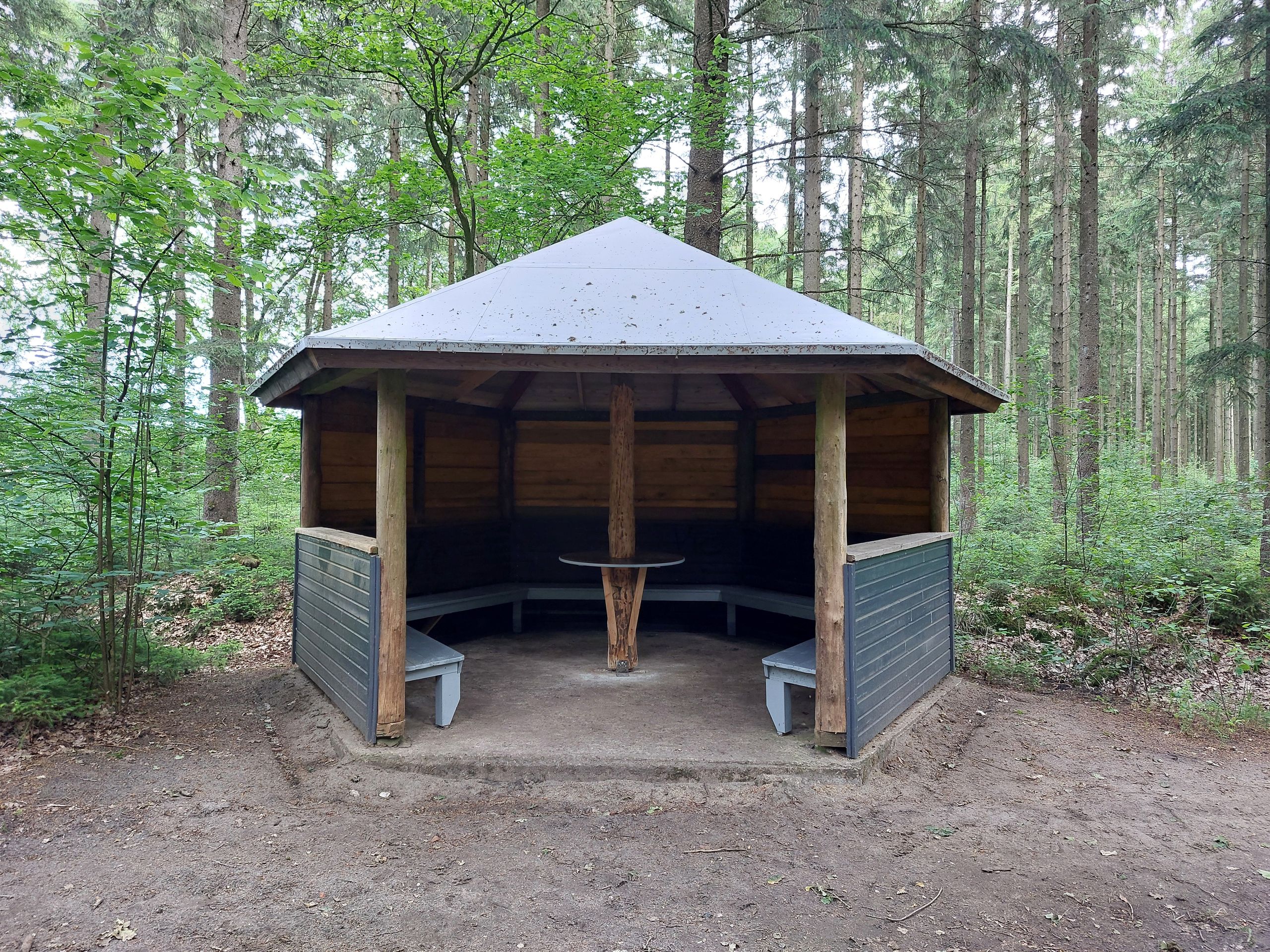 Offener Holzpavillon im Wald mit umlaufenden Bänken und zentralem Tisch unter einem hellen Dach.