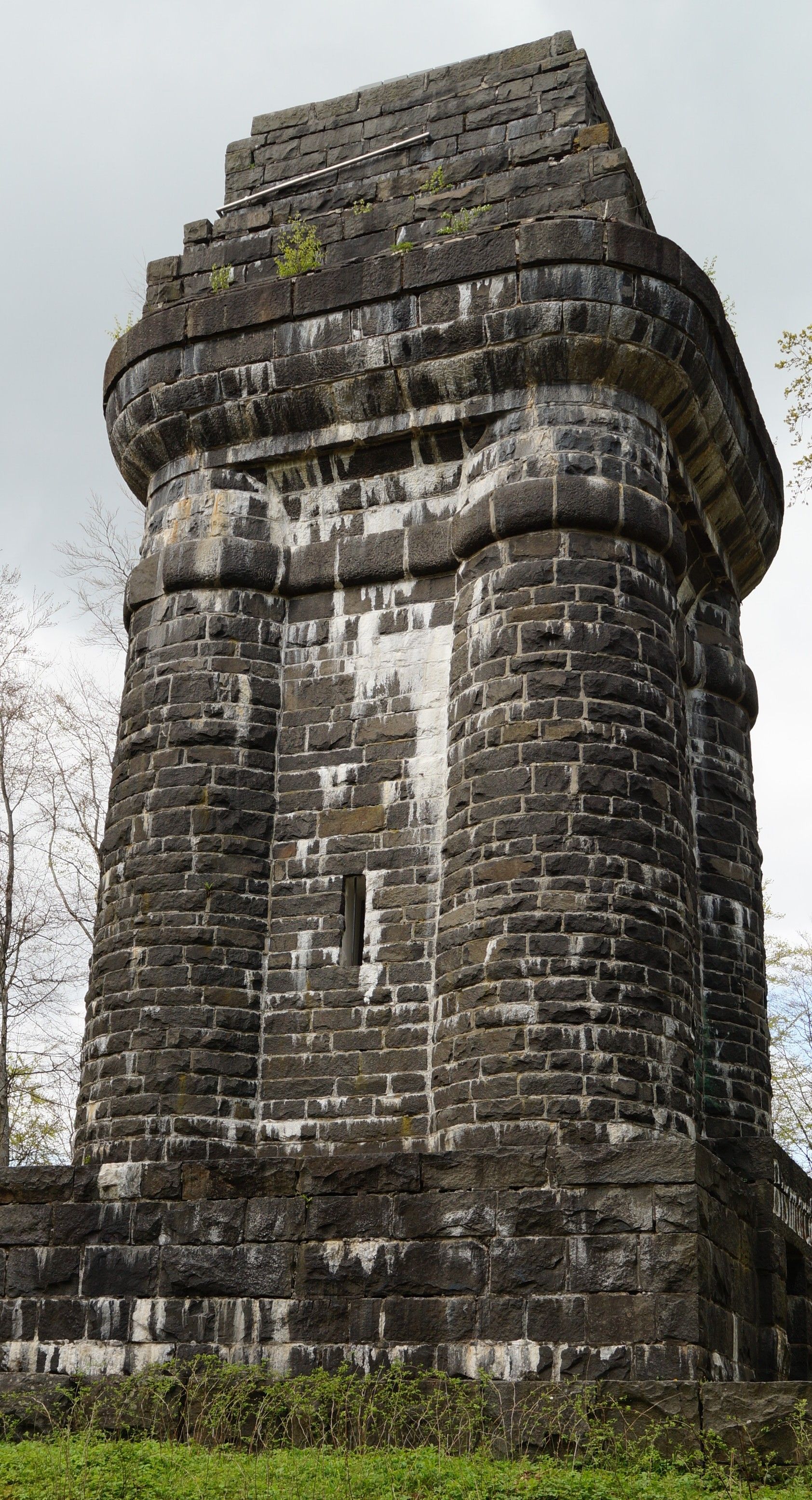 Massiver, verwitterter Steinturm mit dunklem Mauerwerk und kleinen Pflanzen auf der Spitze vor grauem Himmel.