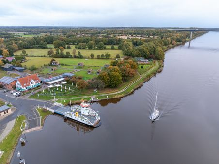 Luftaufnahme des NOK mit einer Fähre und einem Segelboot. Im Hintergrund sind grüne Felder, ein Gebäude mit rotem Dach und mehrere kleine Häuser zu sehen.