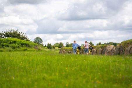 Zwei Personen mit gelben Fahrrädern stehen auf einem grünen Feldweg. Der Himmel ist bewölkt, und die Umgebung ist von üppigem Grün geprägt.