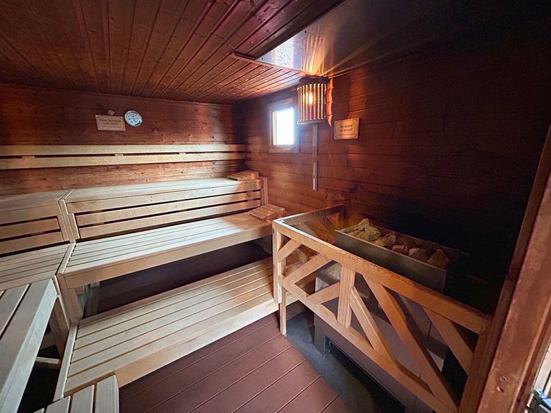 Eine traditionelle Holzsauna mit Holzbänken und einem Ofen. Ein kleines Fenster lässt Licht herein, und ein Thermometer ist an der Wand angebracht.