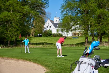 Zwei Golfer auf einem gepflegten Grün vor dem Schloss Breitenburg, umgeben von üppigem Grün und Bäumen.