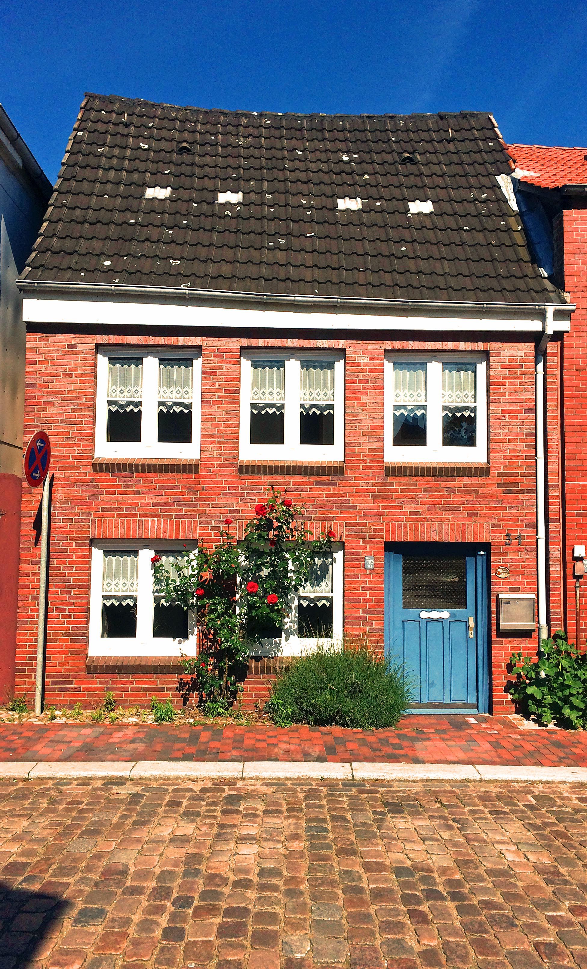 Kleines Backsteinhaus mit blauen Türen und weißen Fenstern. Rote Rosen ranken an der Fassade. Ein gepflasterter Gehweg führt am Haus vorbei.