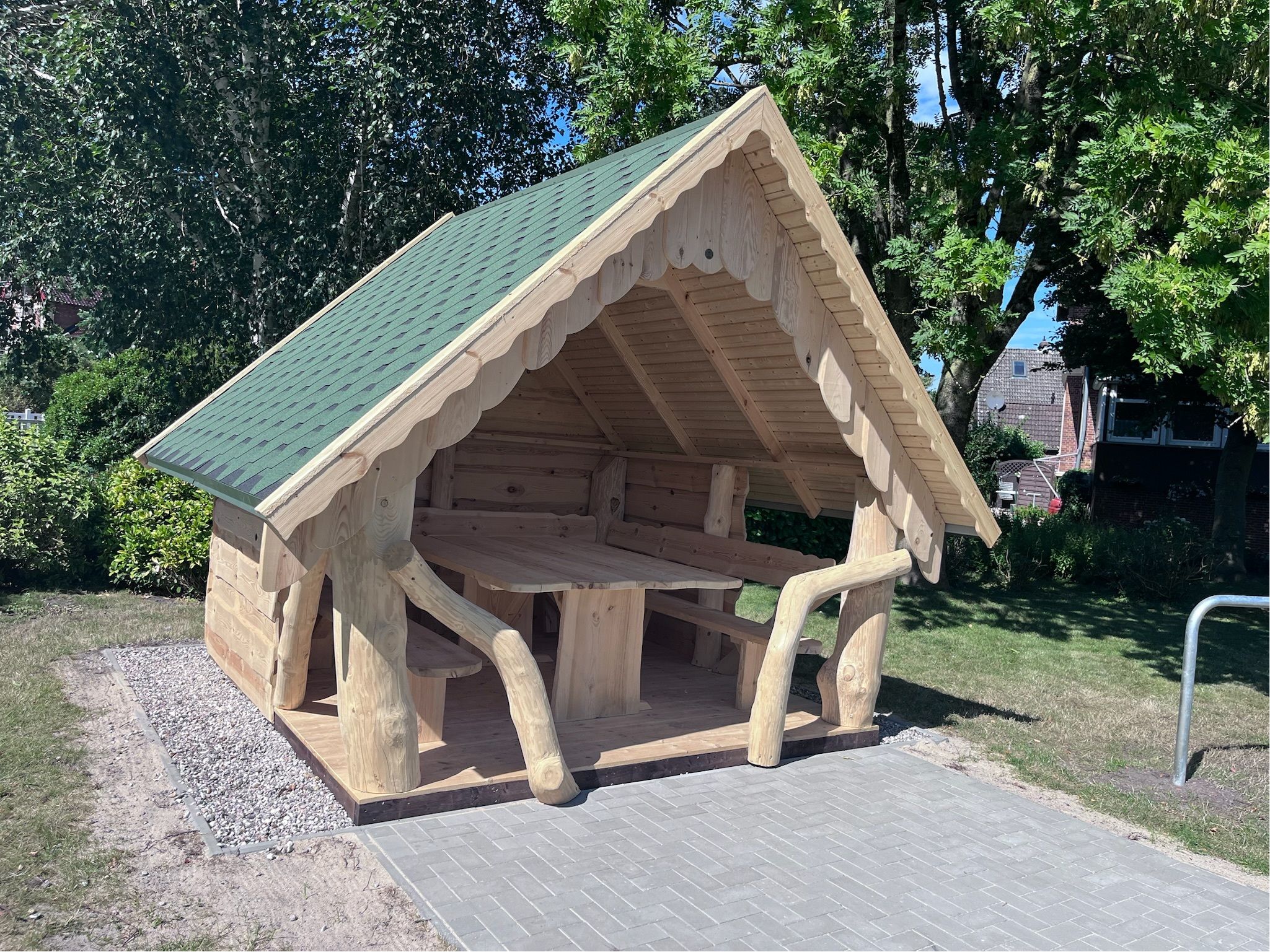 Schutzhütte mit grünem Dach und Sitzbank in einem Park, umgeben von Bäumen und Rasen.