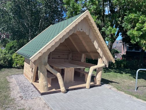 Schutzhütte mit grünem Dach und Sitzbank in einem Park, umgeben von Bäumen und Rasen.