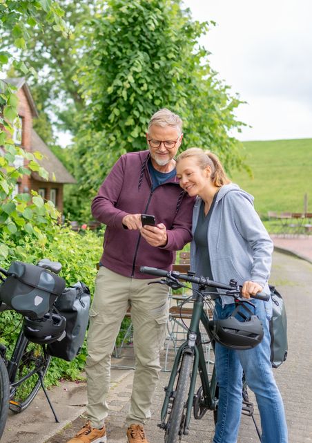 Ein Paar mit Fahrrädern schaut gemeinsam auf ein Smartphone. Sie stehen auf einem Weg, umgeben von grüner Natur.