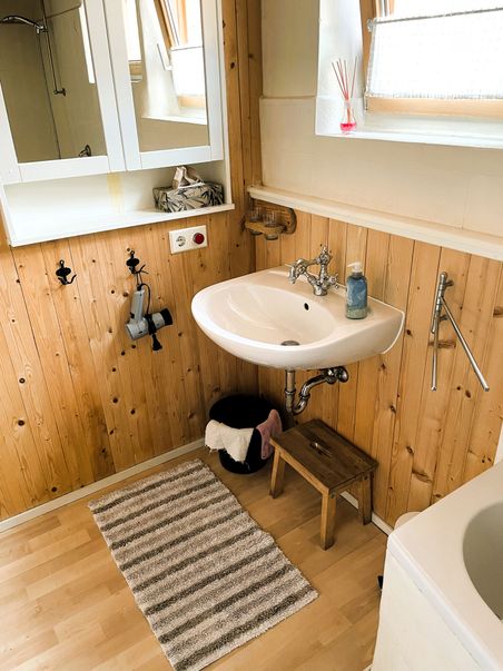Gemütliches Badezimmer mit Holzverkleidung, Waschbecken, Spiegel und kleinem Hocker. Ein gestreifter Teppich liegt auf dem Boden.