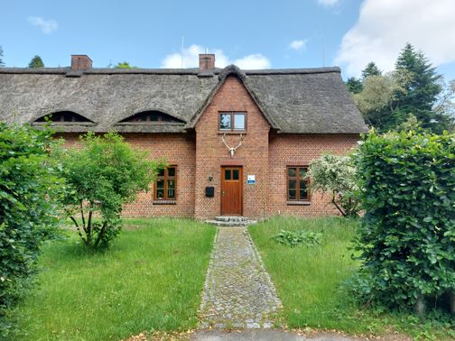 Ein Backsteinhaus mit Reetdach und gepflegtem Gartenweg, umgeben von grünen Bäumen und Sträuchern. Der Himmel ist leicht bewölkt.