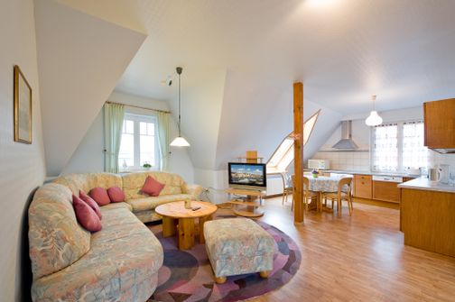 Helles Wohnzimmer mit floralem Ecksofa, rundem Holztisch und Fernseher. Offene Küche mit Esstisch, Holzschränken und Dachfenster.