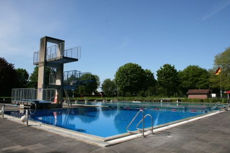 Freibad mit einem Sprungturm und klarem Wasser. Im Hintergrund sind Bäume.
