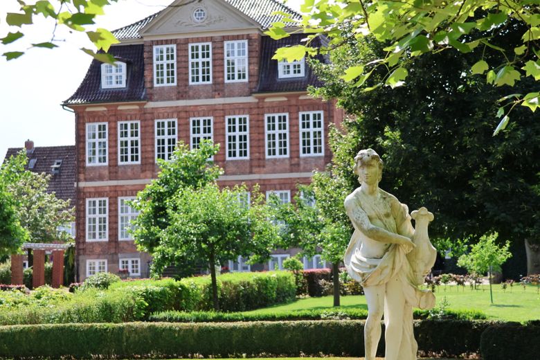 Eine weiße Statue steht in einem gepflegten Garten vor einem historischen Backsteingebäude mit vielen Fenstern. Umgeben von grünen Bäumen und Sträuchern.