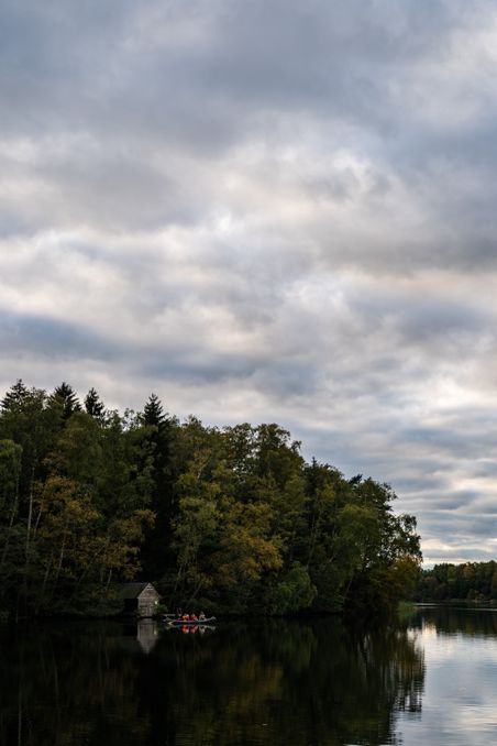 Paddler im Kanu auf dem Pipersee vor bewaldetem Ufer und kleiner Hütte unter bewölktem Himmel.